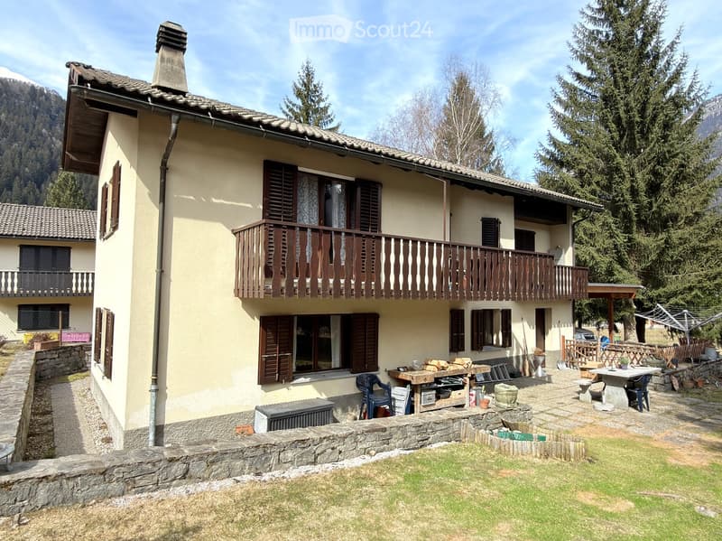 Two-story house with beige walls, wooden shutters, and a balcony on the upper floor. The house is surrounded by a stone wall, a grassy lawn, and a small patio area with outdoor furniture and tools.