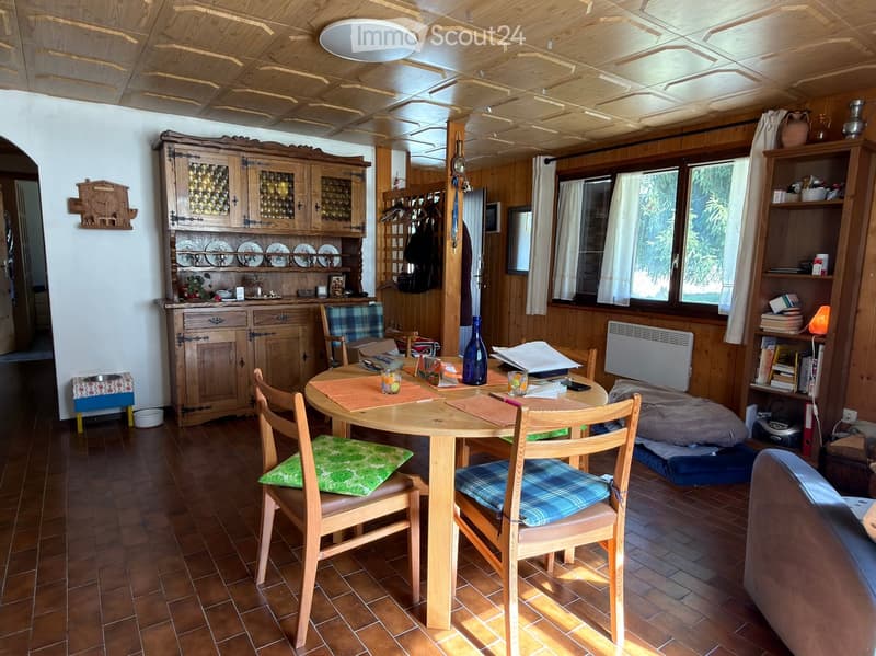 Cozy dining area with wooden table, chairs, and a wooden cupboard. The room features wooden walls, a tiled floor, and large windows with curtains.