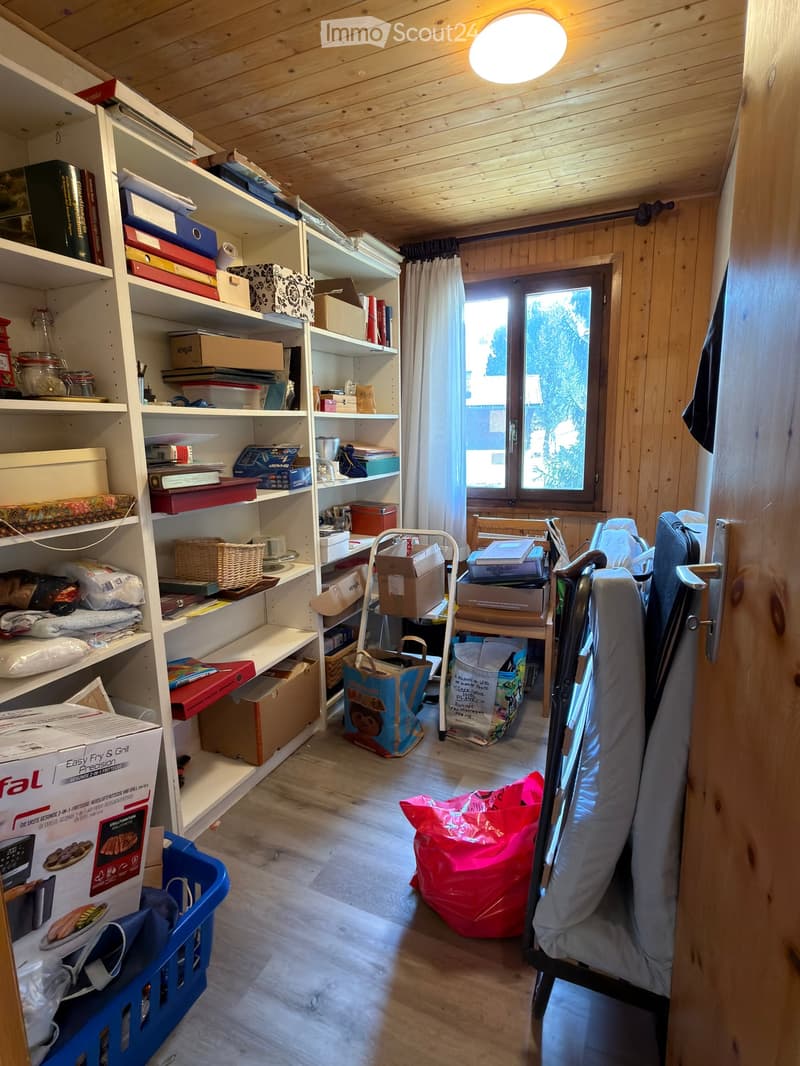 A cluttered room with wooden walls and ceiling, featuring shelves filled with books, boxes, and various items, a window with curtains, and a partially visible sofa and chair.