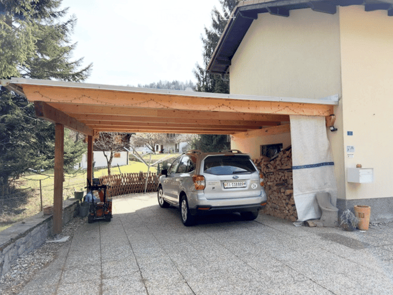 Carport with wooden roof and pillars, silver SUV parked inside, stack of firewood, and a small cart nearby.