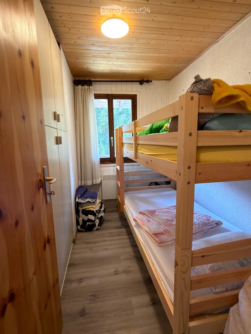 Cozy wooden bedroom with bunk bed, wooden floor, and built-in cabinet. Features a window with curtains and a door leading to another room.