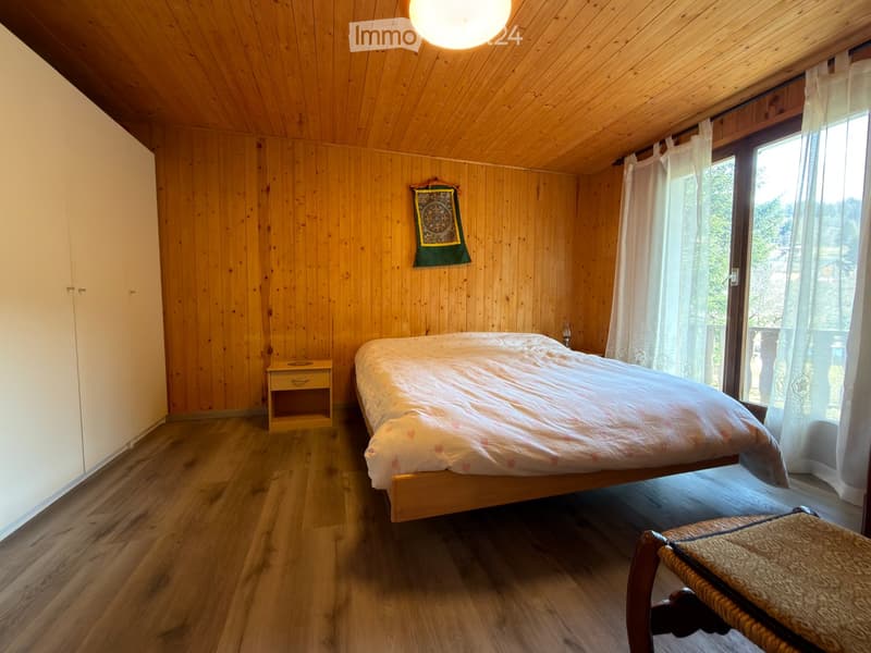 Cozy wooden bedroom with a bed, nightstand, and a small stool. Large window with sheer curtains offers a scenic view.