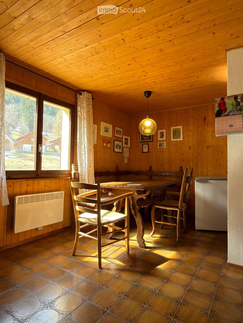 A cozy, wooden-paneled room with a dining table, chairs, a refrigerator, and a radiator. The room features a large window with a view, wooden walls, and a patterned tile floor.