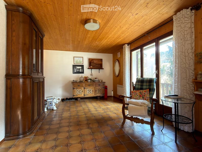 Cozy living room with wooden ceiling, tiled floor, large window with curtains, armchairs, small table, cabinet, and decorative items.
