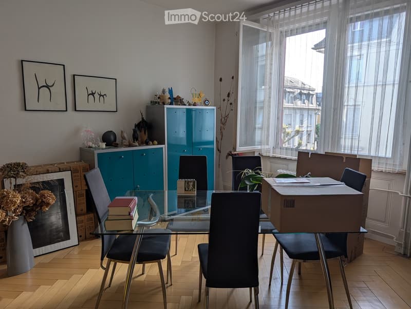 Dining room with a glass table, four chairs, a cabinet, framed pictures, and a vase with dried flowers. The room has wooden flooring and large windows with sheer curtains.