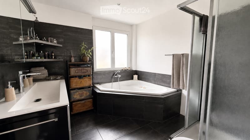 Modern bathroom with a large bathtub, sink, shelves, and a glass shower enclosure. Features dark tiles and a small plant on a wooden crate shelf.