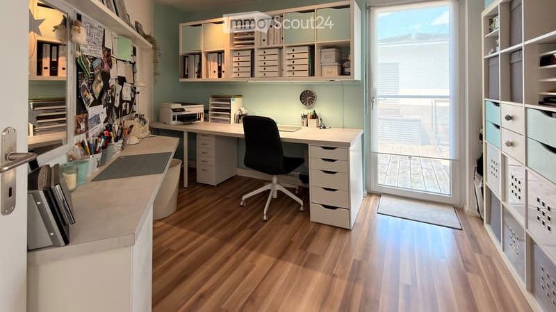 Well-organized home office with a large wooden desk, ergonomic chair, printer, clock, and multiple shelves filled with storage boxes and office supplies. Large window with blinds allows natural light to fill the space.