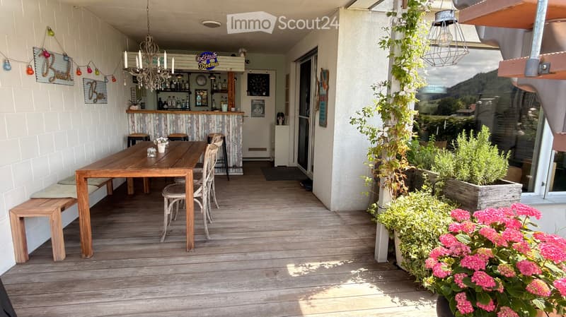 Wooden deck with a dining table, chairs, bench, and bar area with stools and chandelier. Potted plants and flowers line the edge.