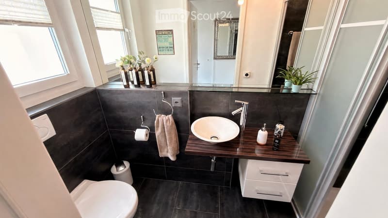 Modern bathroom with dark tiles, a wooden vanity, a sink, a toilet, a towel, and a mirror. There are decorative bottles and a small plant on the windowsill.
