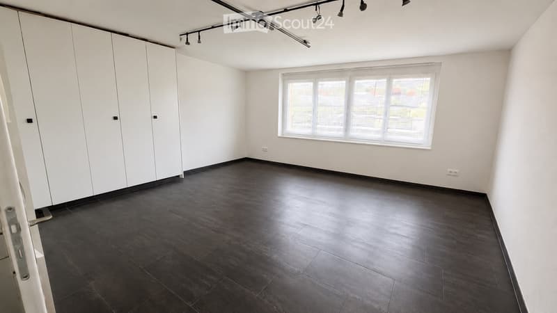 Empty room with white walls, dark wooden flooring, large window with blinds, and built-in white closet units.