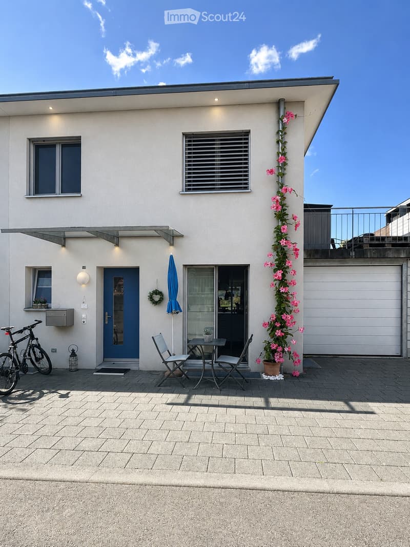 Modern house exterior with a paved patio area featuring a small table, two chairs, a bicycle, and a potted plant with pink flowers. The house has a blue front door, large windows, and a garage.