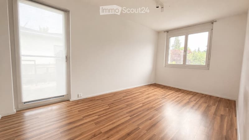 Empty room with wooden flooring, white walls, large window with blinds, and a sliding glass door leading to a balcony.