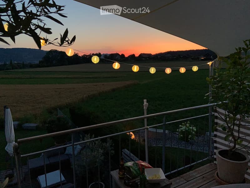 Balcony with a scenic view of a field and mountains at sunset, decorated with string lights, a small table with a bowl of snacks and drinks, and potted plants.