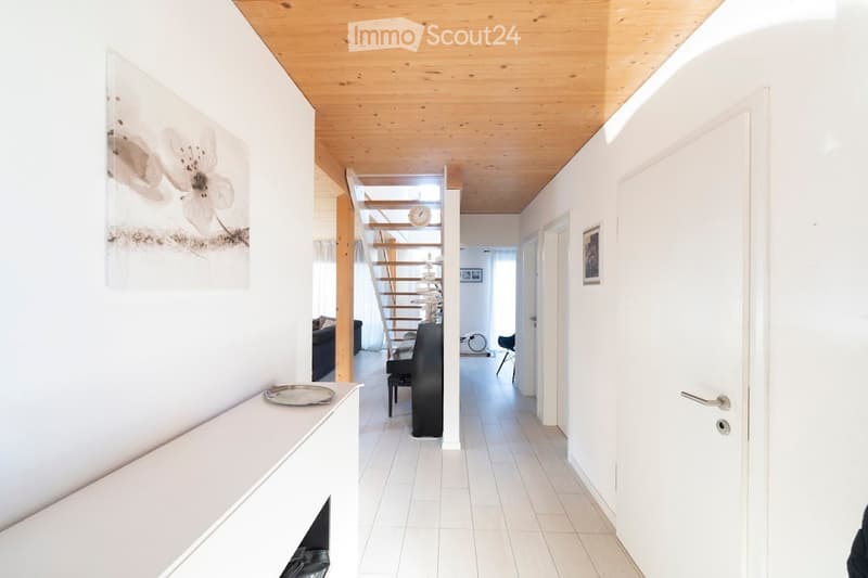 Bright hallway with white walls, wooden ceiling, and tiled floor. Features a staircase leading upwards, a small console table with a decorative tray, and a framed artwork on the wall.