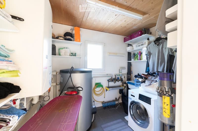 Utility room with wooden ceiling, white walls, washing machine, ironing board, shelves with folded towels, storage bins, and a small window.