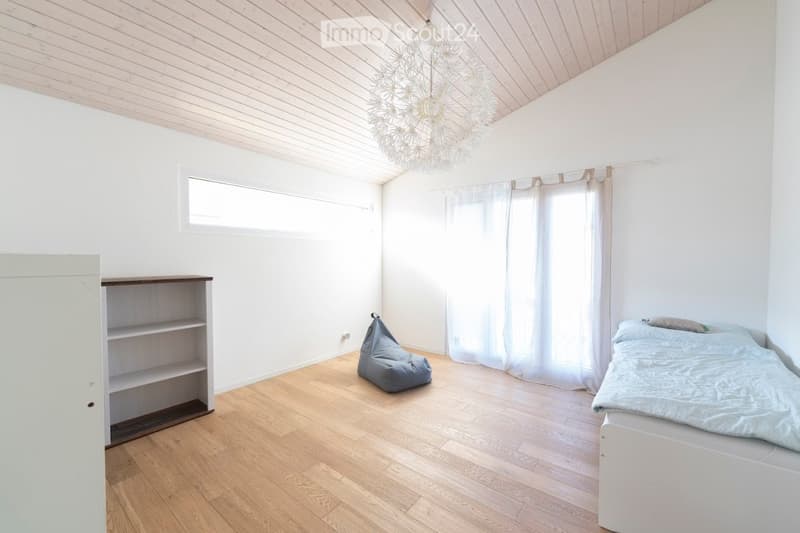 Bright, minimalist bedroom with wooden flooring, white walls, and a slanted wooden ceiling. Features a single bed with light blue bedding, a bean bag chair, a small shelf, and sheer white curtains on the windows.