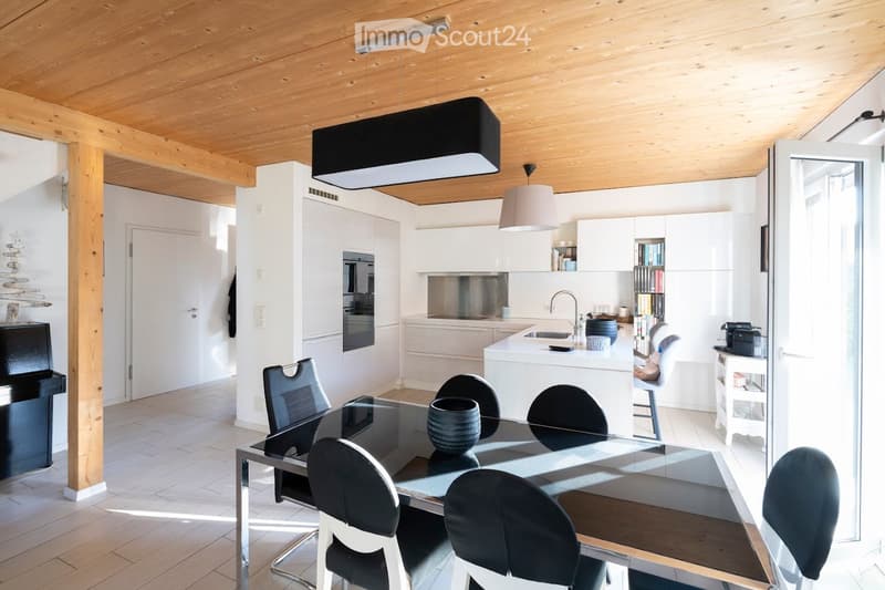 Modern kitchen and dining area with white cabinets, a central island, and a glass dining table surrounded by chairs. The space features wooden beams on the ceiling and a sleek, contemporary design.