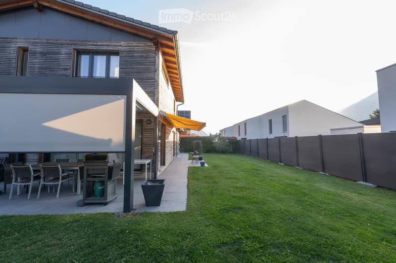 Well-maintained garden with a covered patio area, dining table, chairs, and a barbecue. The patio is adjacent to a modern house with wooden siding and large windows. The garden is enclosed by a wooden fence and features a neatly trimmed lawn.