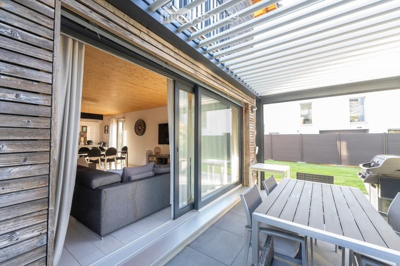 Modern covered balcony with wooden decking, glass sliding doors leading to a living space, outdoor dining table and chairs, and a view of a well-maintained garden.