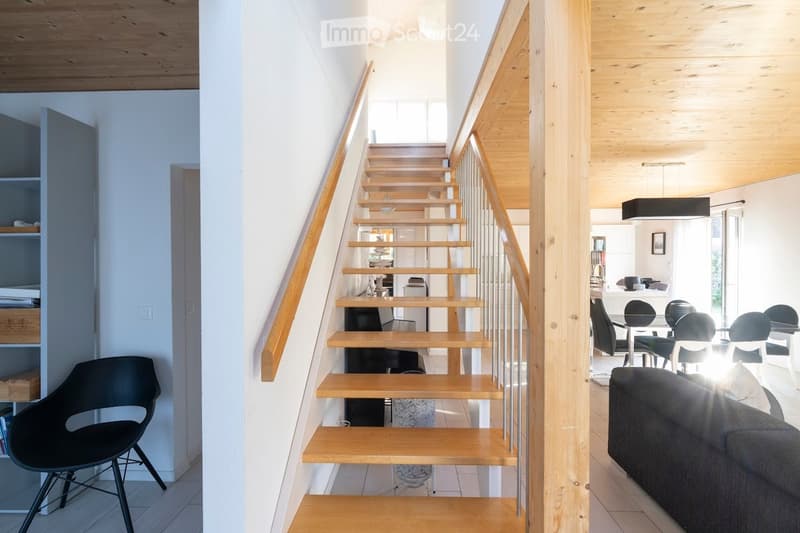 Modern hallway with wooden staircase, white walls, and visible rooms including a living area with a sofa and dining area with chairs and table.