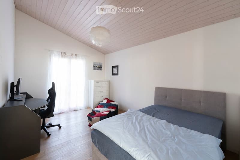 Bright bedroom with a double bed, desk with a computer, chair, and a small cabinet. The room features wooden flooring, white walls, and a large window with sheer curtains.