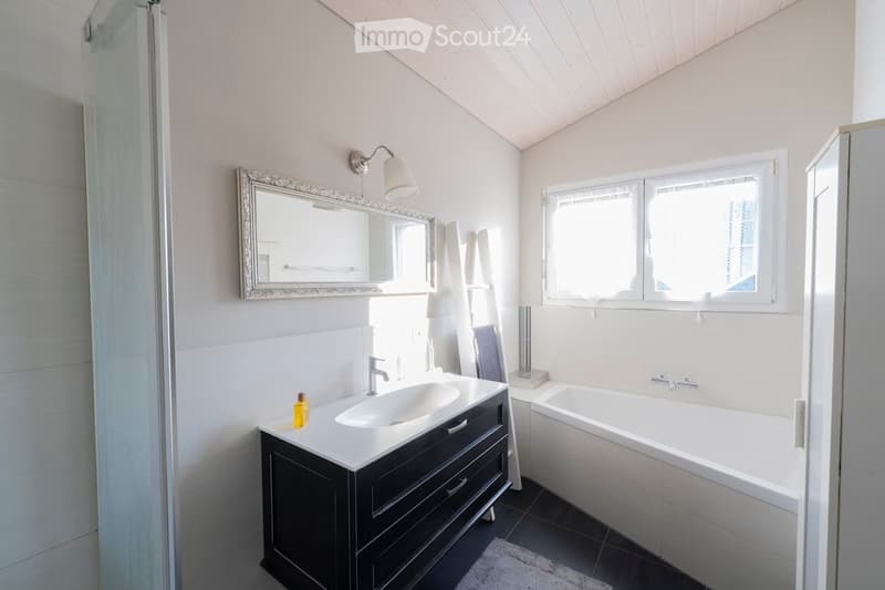 Modern bathroom with a dark vanity, white sink, bathtub, and a large mirror with a decorative frame. The room features a window and a towel hanging on a rack.