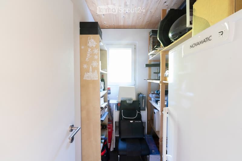 A small, furnished room with wooden shelves, a black chair, a refrigerator, and a lamp. The room has a slanted wooden ceiling and a window letting in natural light.