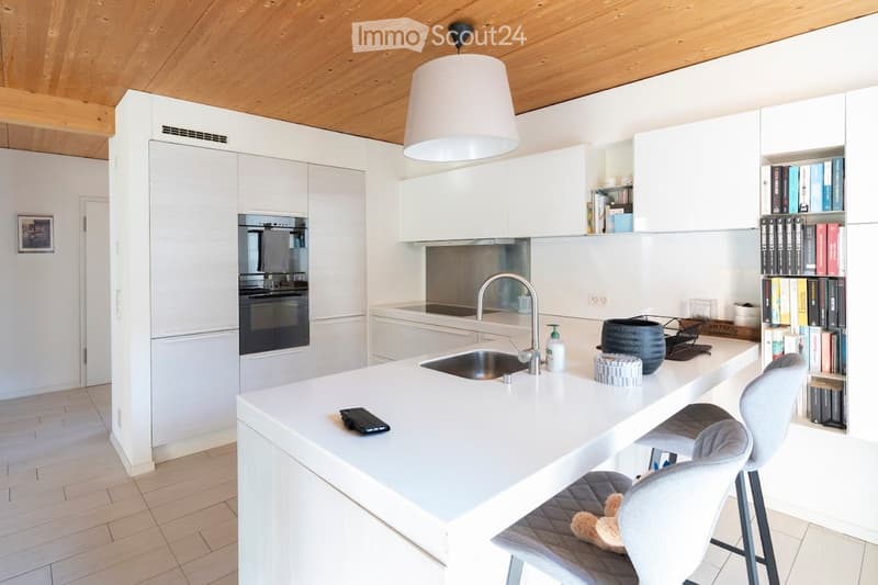Modern kitchen with white cabinets, a central island, and a sink. Features include a built-in oven, a sleek faucet, and a bookshelf with books and decorative items.