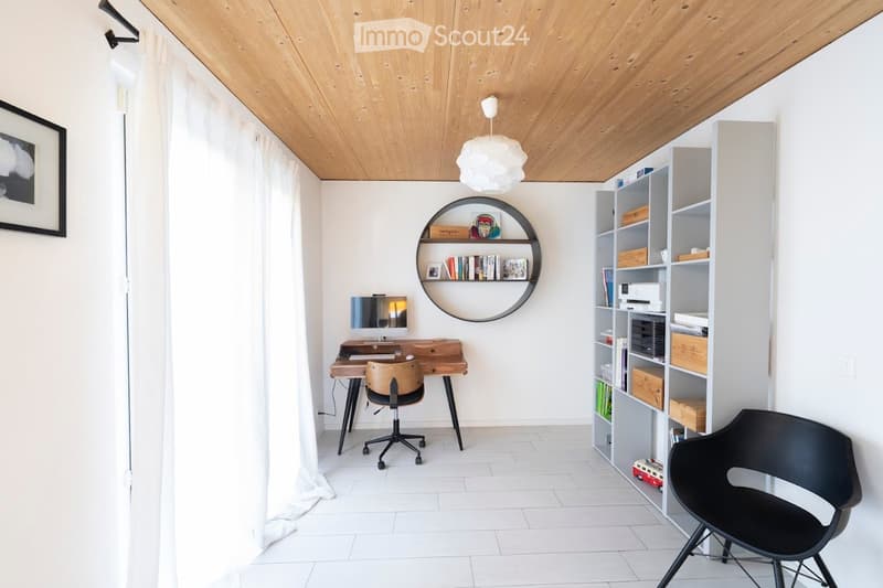 A modern, well-lit room with a wooden ceiling, white walls, and a large window with white curtains. It features a wooden desk with a computer monitor, a black office chair, a round wall shelf with books and decorative items, and a white shelving unit with