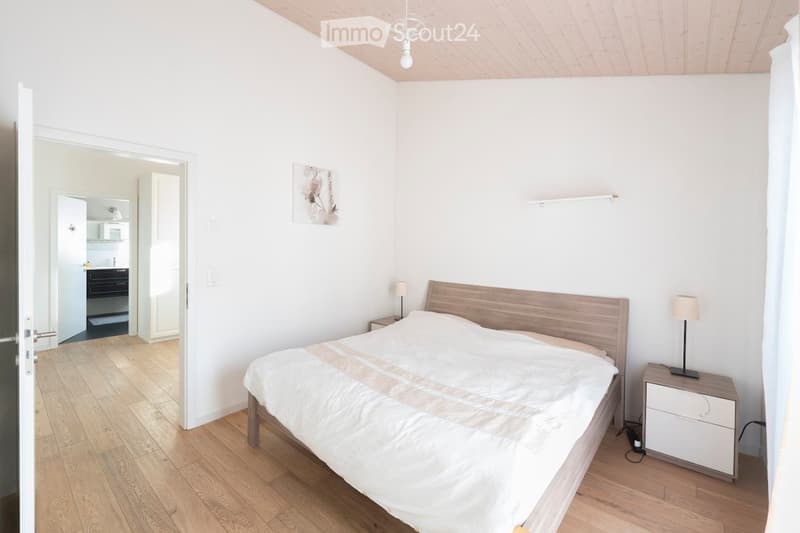 Modern bedroom with a wooden bed, white bedding, nightstands, and lamps. The room features light wooden flooring and white walls, with a small adjacent bathroom visible through an open doorway.