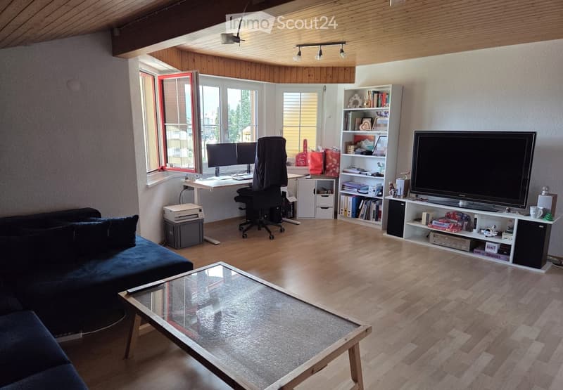 Modern living room with wooden floors, large windows, a dark blue sofa, a glass coffee table, a desk with a chair, a bookshelf, and a television on a stand.