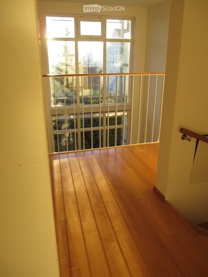 A narrow hallway with wooden flooring and a metal railing leading to a window with a view of the outside. The walls are painted a light color, and the space is well-lit with natural light coming through the window.