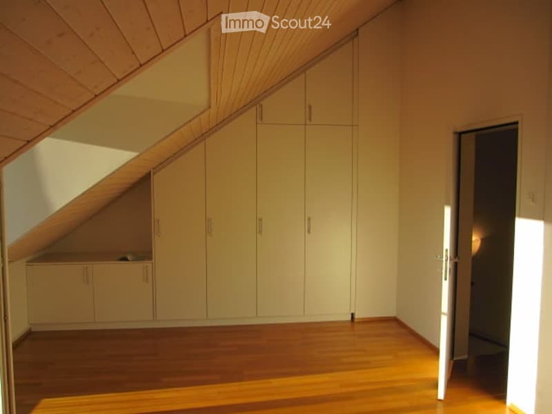 Empty attic room with wooden floor, white built-in wardrobes, and slanted wooden ceiling.