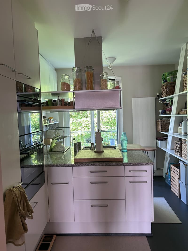 Modern kitchen with light pink cabinets, granite countertops, and stainless steel appliances. Features include a sink with a faucet, an oven, a cutting board, and various jars and baskets on shelves.