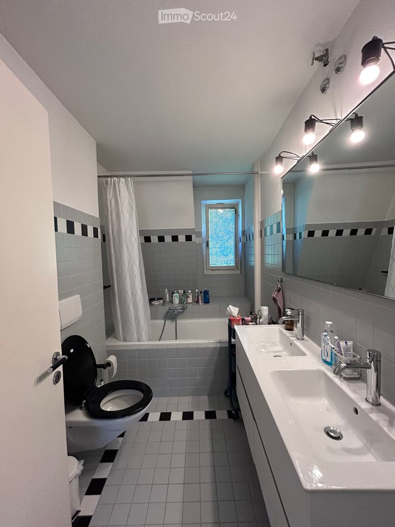 Modern bathroom with a toilet, double sink, bathtub, and large mirror. Features tiled walls and floor, a window with blinds, and multiple light fixtures.