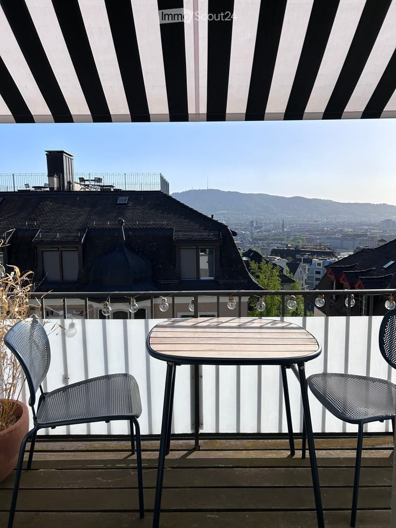 Balcony with a small table and two chairs, overlooking a cityscape with mountains in the background.