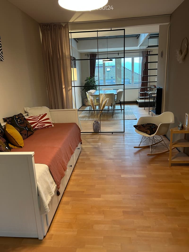 A modern, open-concept room with a daybed, a rocking chair, a dining area with a table and chairs, and a view of a rooftop terrace.