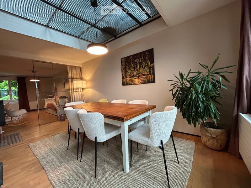 Dining room with a wooden table, white chairs, large plant, mirror, and artwork on the wall.