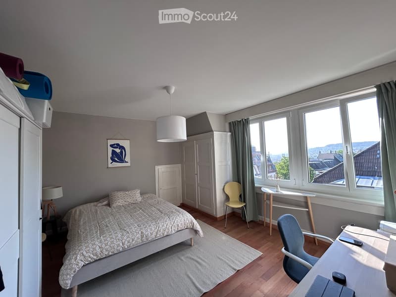 Cozy bedroom with a bed, desk, chair, and large windows offering a city view.