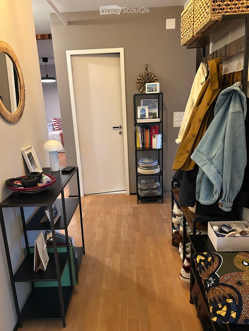 A well-lit hallway with wooden flooring, featuring a coat rack with jackets, a shelf with books and decorative items, and a small table with a lamp and decorative bowl.