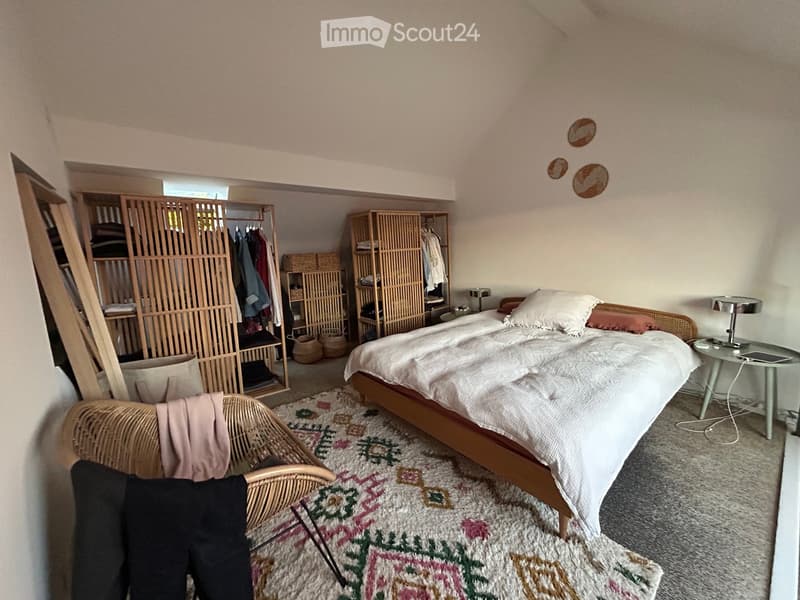 Cozy bedroom with a wooden bed, nightstand, wicker chair, and open wooden wardrobe with shelves and hanging space.