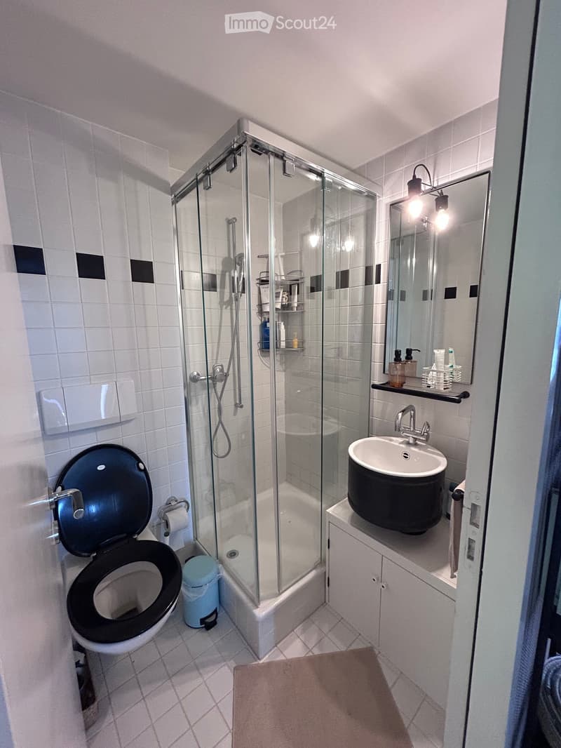 Modern bathroom with white tiles, a black toilet, a sink with a modern faucet, a glass-enclosed shower, a mirror with built-in lighting, and a small shelf holding toiletries.