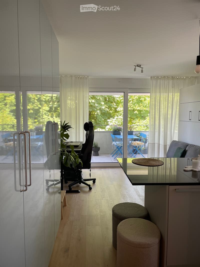 Modern living space with light wooden flooring, large windows, and a balcony area with outdoor furniture.