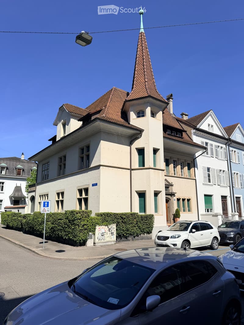 A multi-story residential building with a distinctive tower and spire, surrounded by other houses and parked cars.