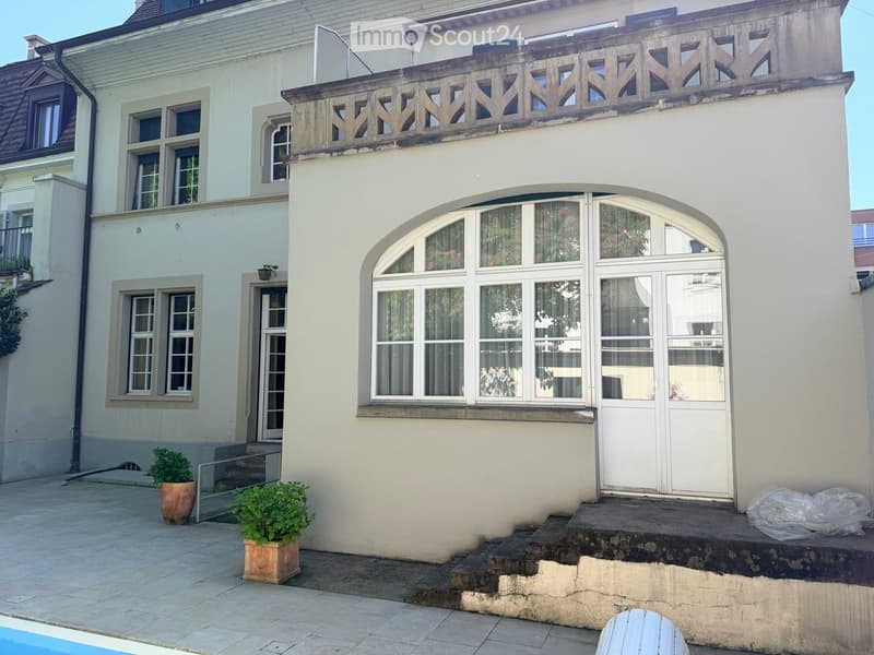 Two-story residential building with white facade, arched entrance, multiple windows, decorative balcony railing, and small garden area with potted plants.