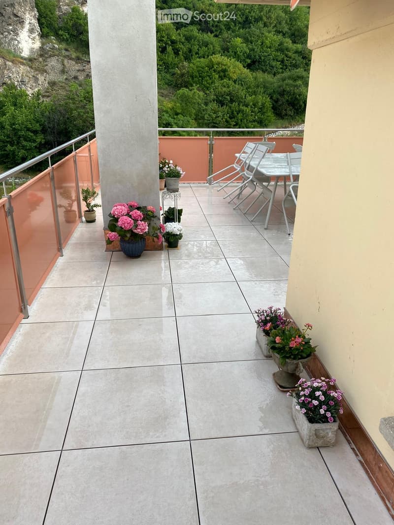 Balcony with tiled floor, metal railing, and outdoor furniture including a table and chairs, surrounded by potted plants and a scenic view of greenery.