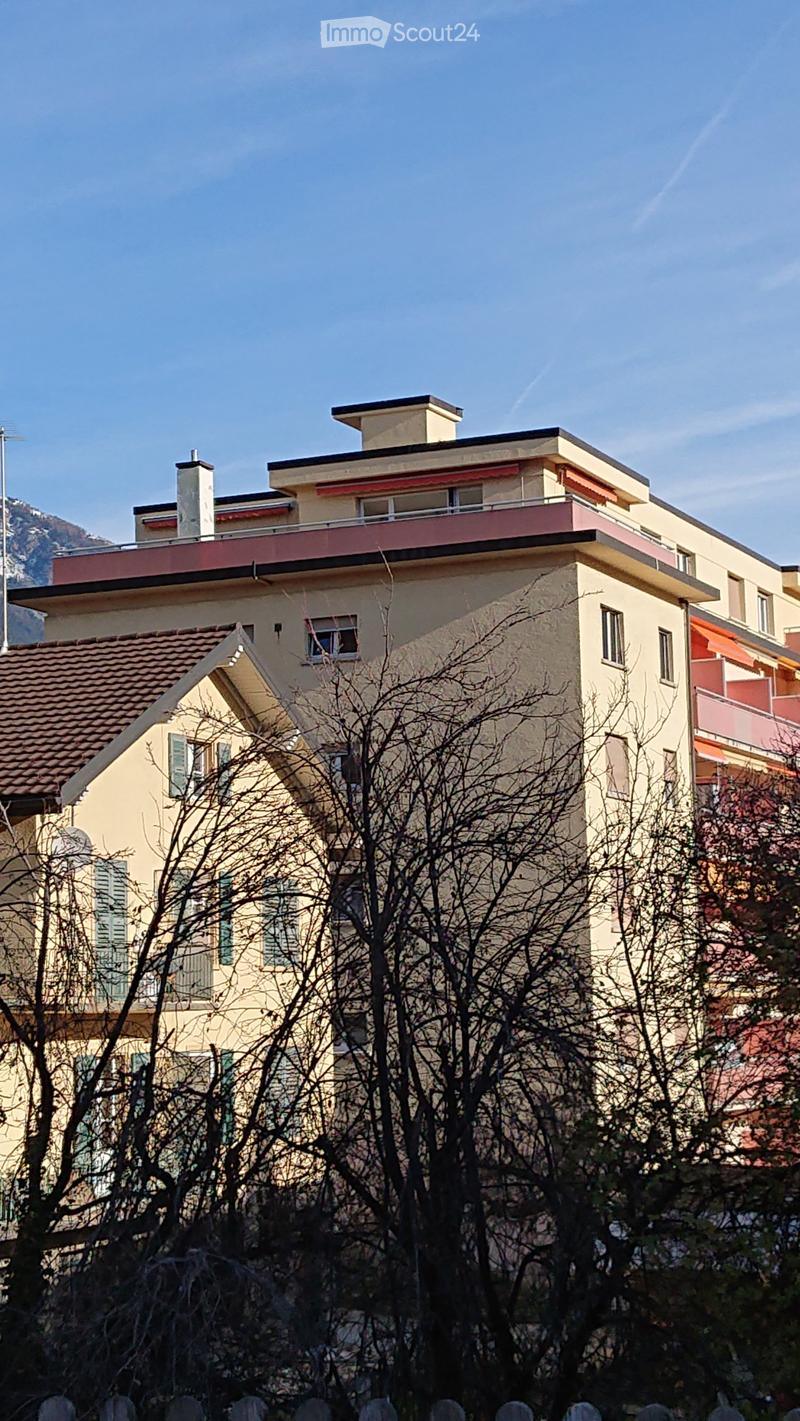 Multi-story residential building with beige walls, red accents, and multiple windows, partially obscured by bare trees.