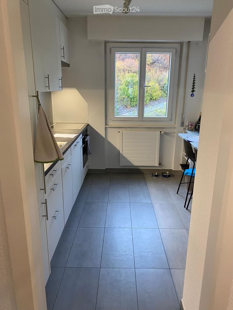 Modern kitchen with white cabinets, tiled floor, and a window overlooking a garden. Includes an oven, sink, and a small table with a chair.