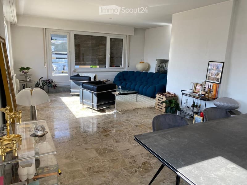 Modern living room with marble flooring, large windows, blue sofa, black leather chair, glass coffee table, and decorative items including a chessboard and framed pictures.