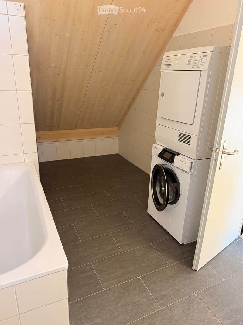 Bathroom with a washing machine and dryer stacked, a bathtub, tiled walls, and wooden slanted ceiling.
